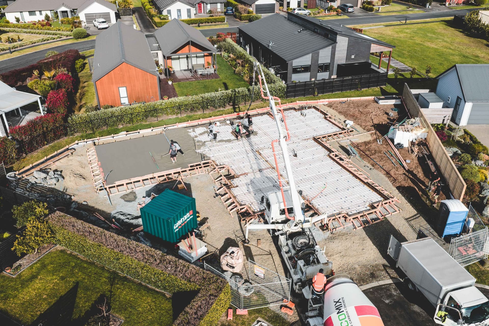 Aerial view of the slab pour on a new home in a Mangawhai Heads cul-de-sac, with a concrete boom pump over the reinforcing steel and the crew screeding the first section
